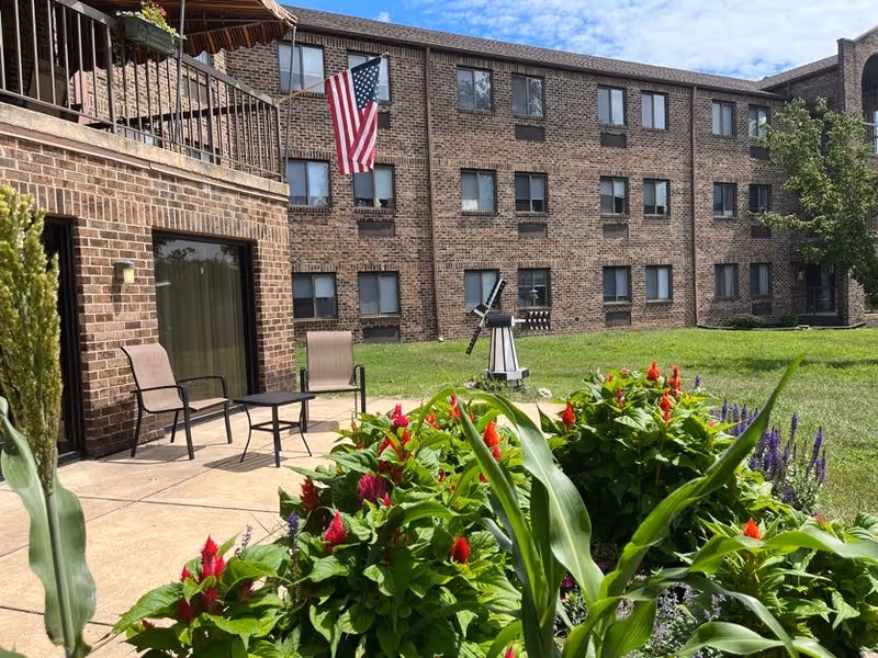 Outdoor patio area with two chairs and a small table next to a brick building. There is a garden with green plants and red flowers in the foreground, and an American flag is hanging on the building. The sky is partly cloudy.