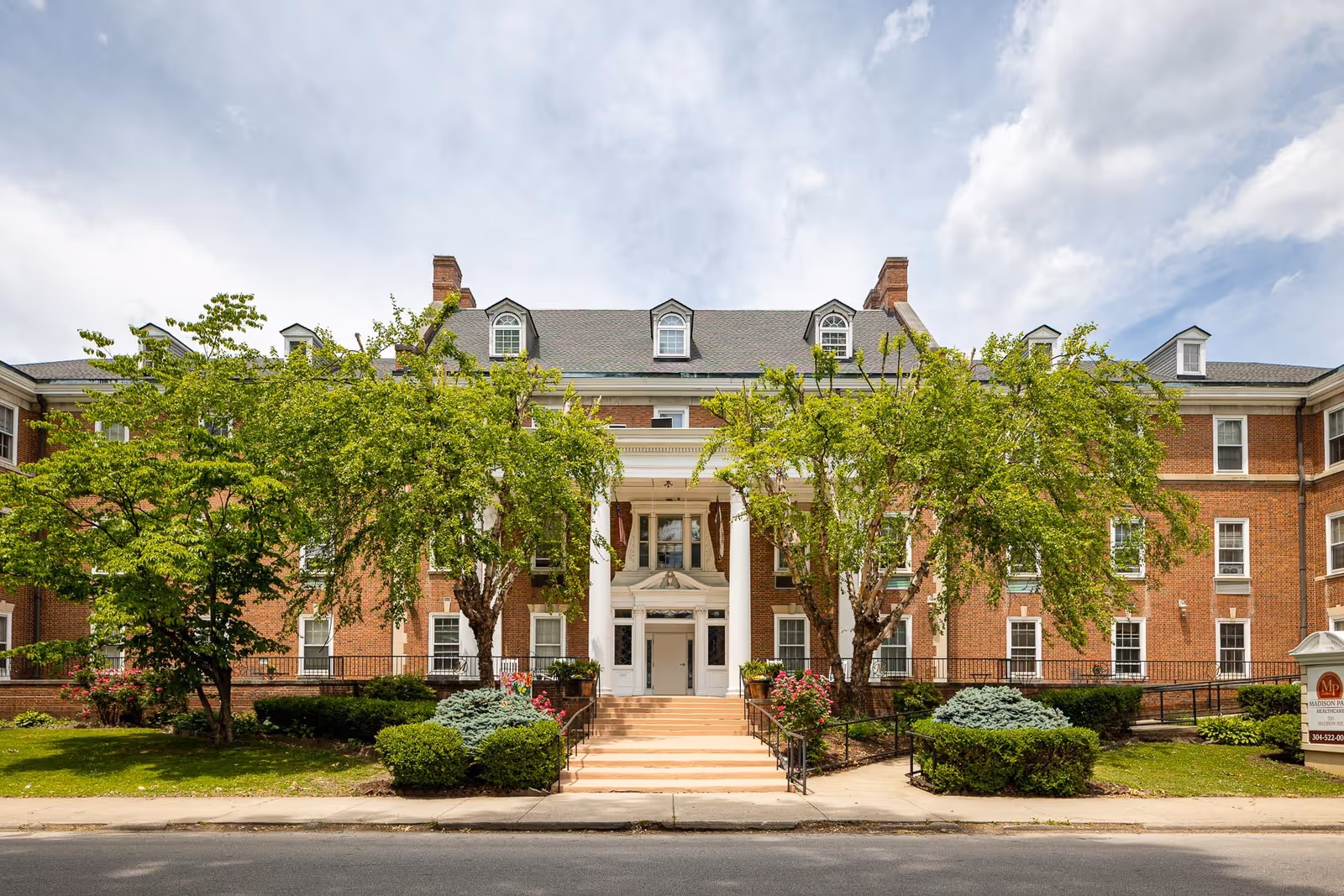 Front exterior of a red-brick, three-story building with white columns, entry steps, and landscaped trees and shrubs.