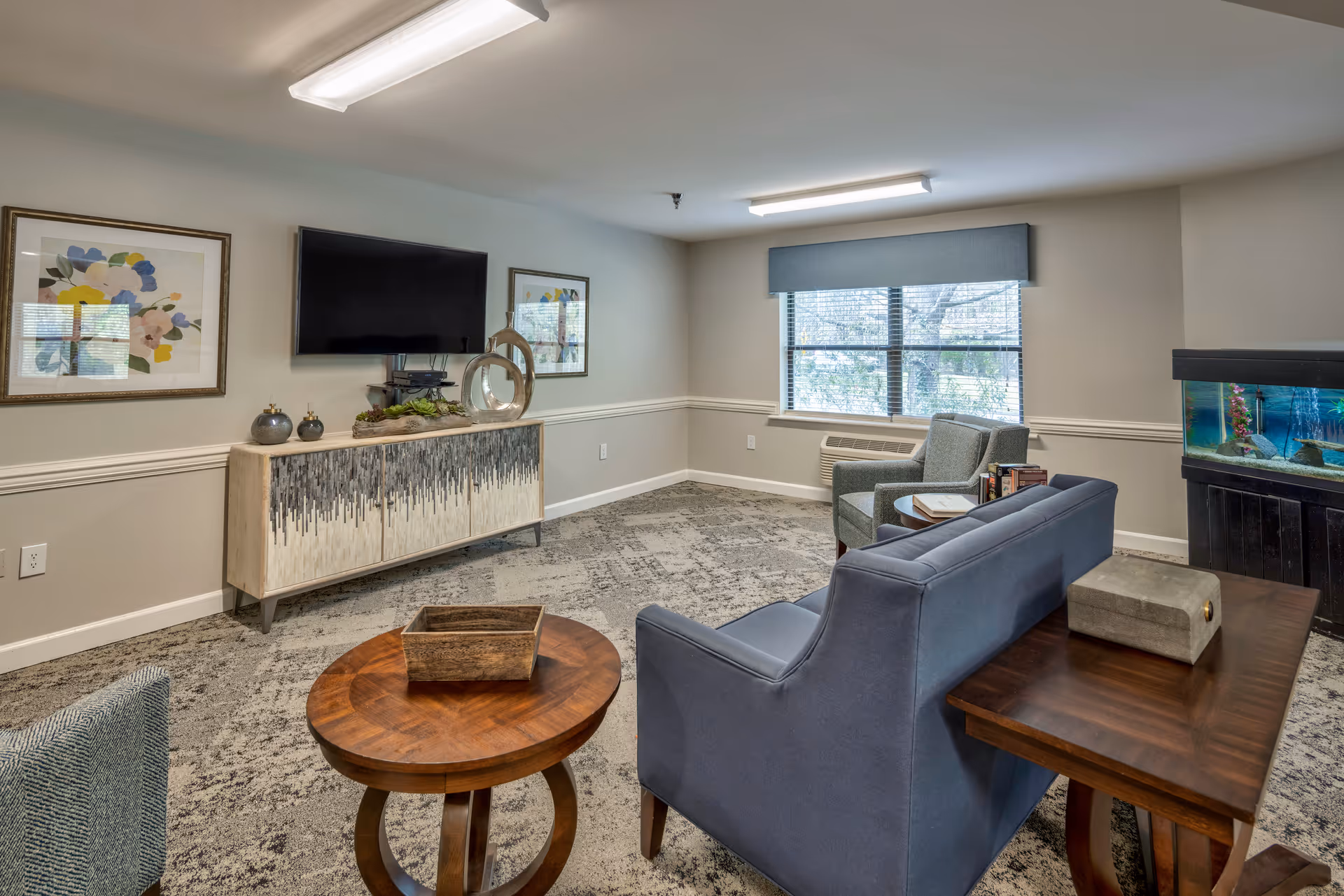 Communal living room with a sofa and chairs facing a wall-mounted TV, decorative cabinet and an aquarium.