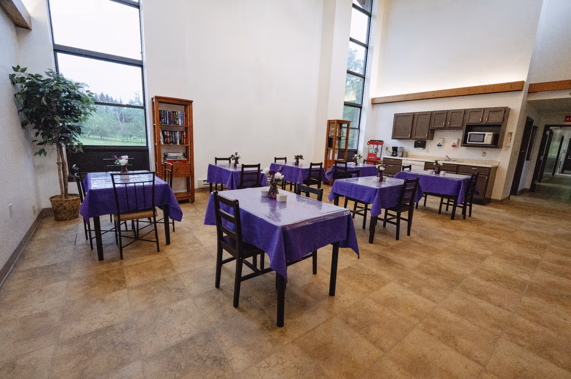 A dining area with several tables covered in purple tablecloths, each with a small flower vase centerpiece. The room has large windows letting in natural light, wooden cabinets with a microwave and coffee maker, and a popcorn machine in the corner. There are bookshelves and a potted plant near the windows.