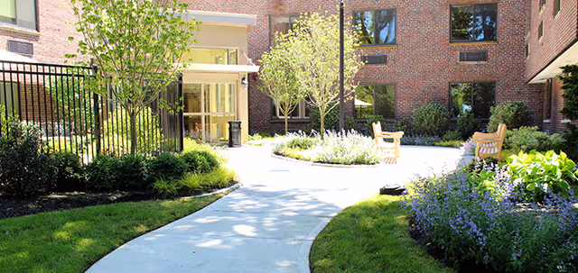 Outdoor courtyard area with a curved concrete pathway, green grass, flowering plants, small trees, and wooden benches. The courtyard is surrounded by a brick building with multiple windows.