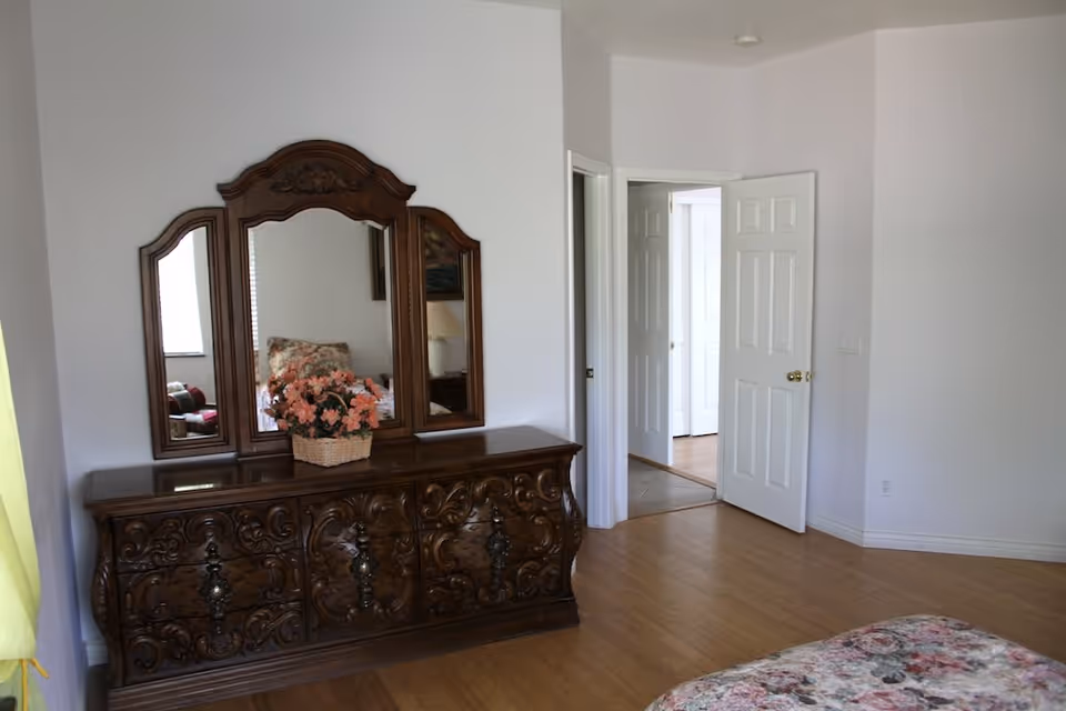 A bedroom with a wooden dresser featuring ornate carvings and a three-panel mirror. On top of the dresser is a basket with pink flowers. The room has light-colored walls and wooden flooring. A bed with a floral bedspread is partially visible in the foreground. Two white doors are open, leading to other rooms.