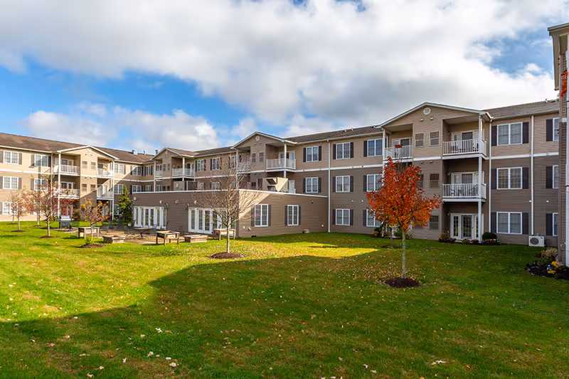 Three-story beige senior living building with balconies overlooking a grassy courtyard with benches and small trees under a partly cloudy sky.