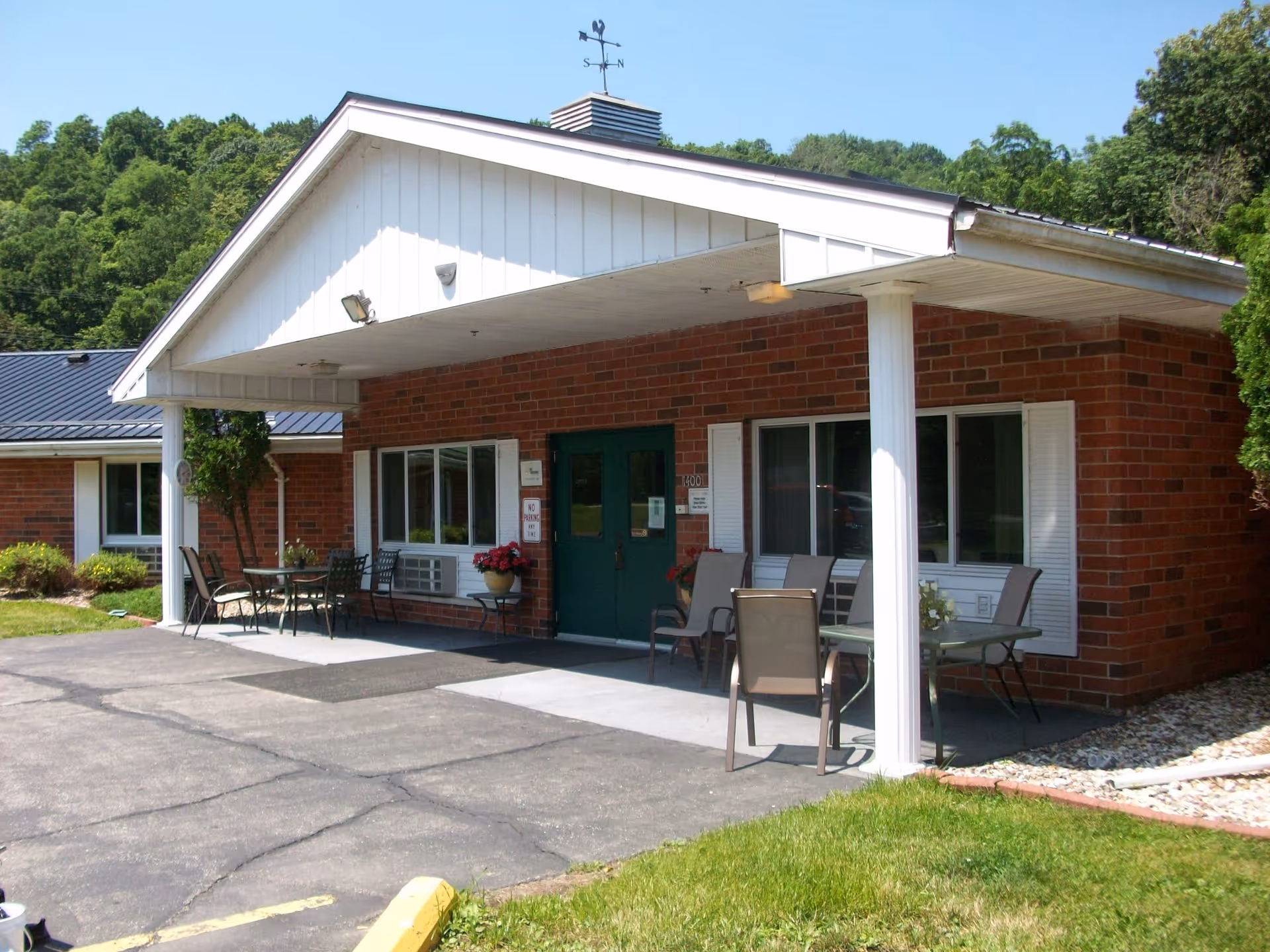 Exterior view of a single-story brick building with a covered entrance supported by white columns. There are green double doors, windows with white shutters, and outdoor seating with tables and chairs on the concrete porch. The building is surrounded by greenery and trees under a clear blue sky.