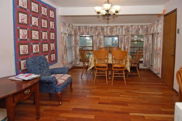 A dining area with a wooden dining table covered with a floral tablecloth and six wooden chairs. The room has hardwood flooring and large windows with floral curtains. To the left, there is a blue upholstered armchair with a quilted cushion and a wooden side table with magazines on it. A colorful quilt with handprint designs hangs on the wall above the armchair. A chandelier with multiple lights hangs from the ceiling.