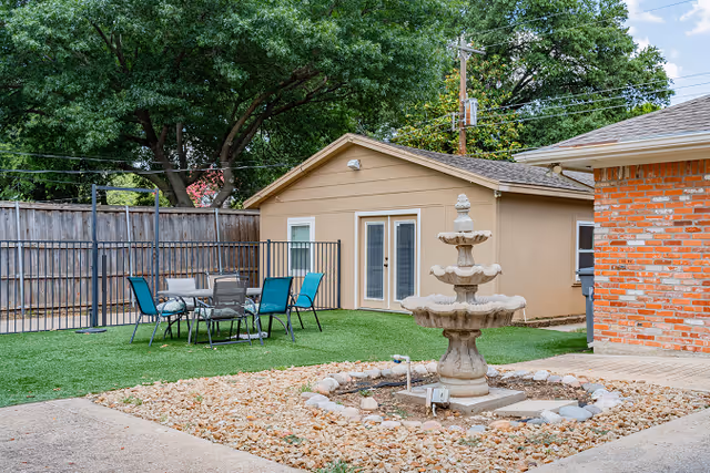 Courtyard with a three-tier stone fountain, patio table and chairs on artificial turf in front of a small beige outbuilding.