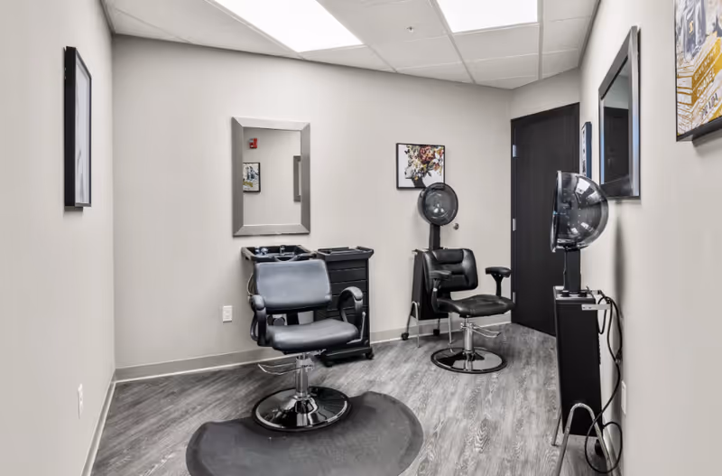 Interior of a small salon room with two black salon chairs, a hair dryer hood, a mirror on the wall, and a small black rolling cart. The room has light gray walls, wood-patterned flooring, and framed artwork on the walls.
