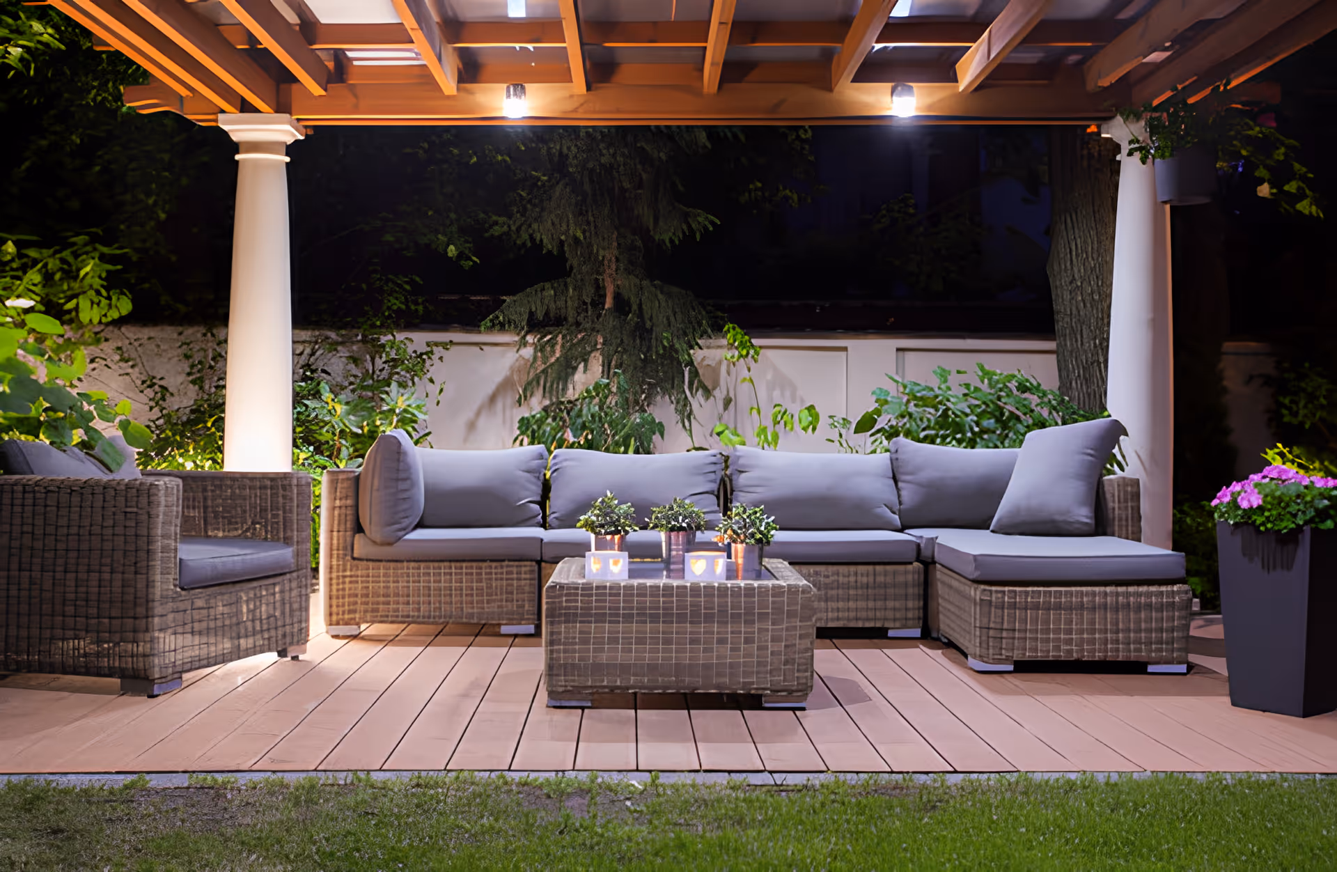 Outdoor patio area at night with a wicker sectional sofa and armchair with gray cushions under a wooden pergola. A wicker coffee table with small potted plants is in front of the sofa. The area is surrounded by greenery and illuminated by overhead lights.