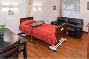 A nursing facility room featuring a hospital-style bed with a red blanket, an overbed table, a black leather couch, a wooden chair and table, a potted plant, and a window with blinds letting in natural light.