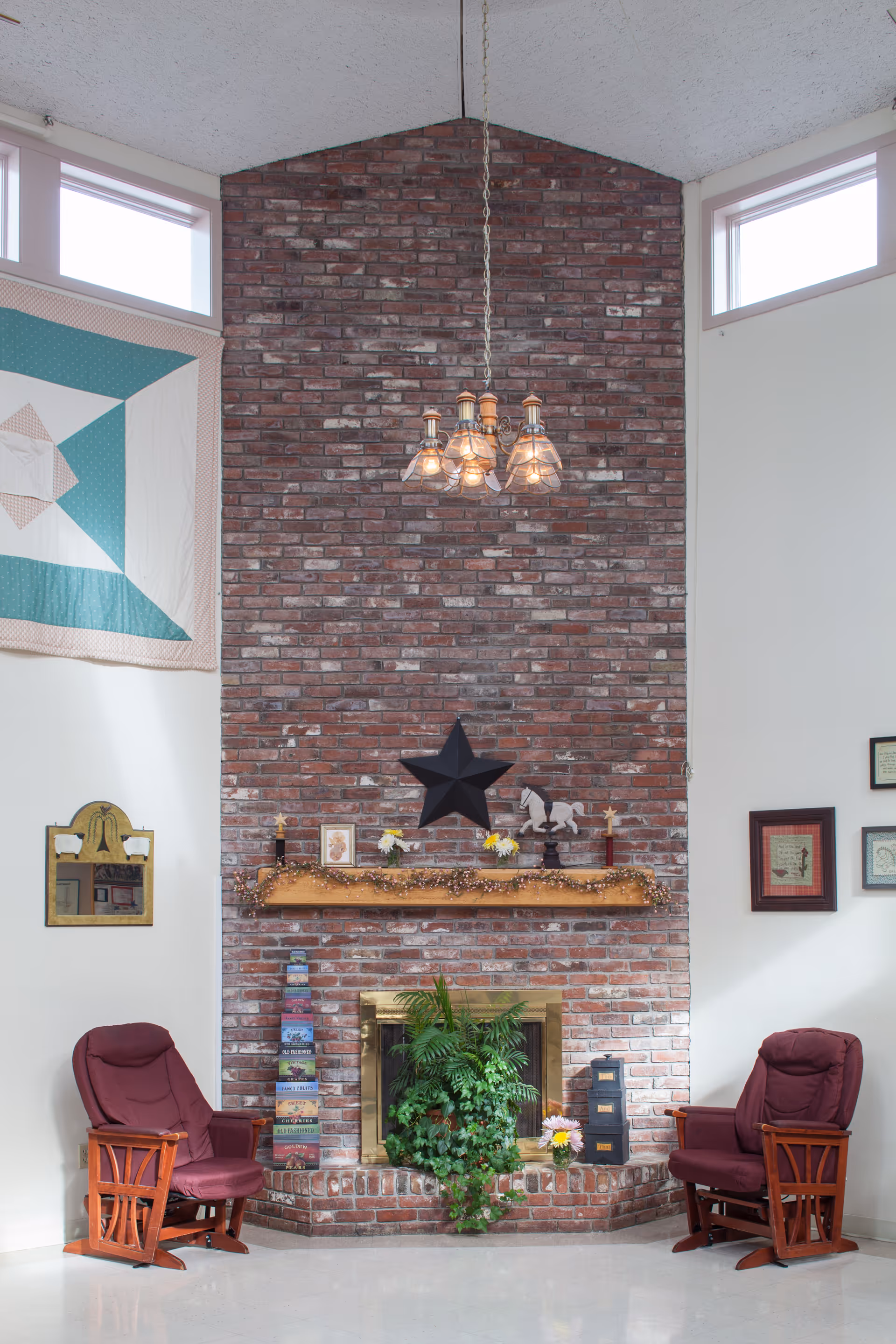 A cozy living room area with a tall brick fireplace as the focal point. The fireplace has a wooden mantel decorated with small framed pictures, flowers, and a black star wall ornament. Two maroon cushioned wooden rocking chairs are positioned on either side of the fireplace. A green leafy plant sits in front of the fireplace. The walls are white with a quilt hanging on the left side and framed pictures on the right. A chandelier with five lights hangs from the ceiling above the fireplace.