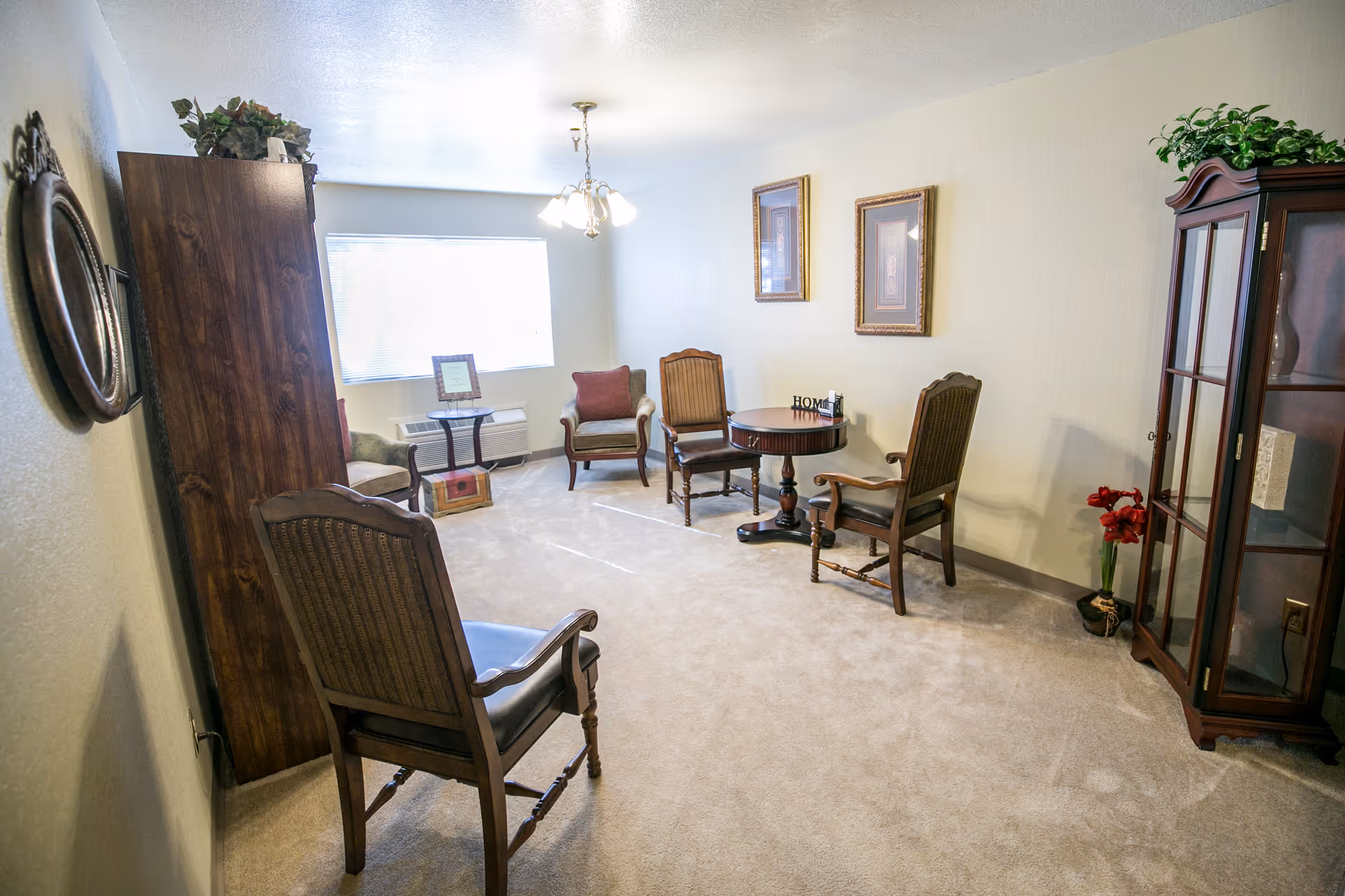 A cozy living room with beige carpet and cream-colored walls. The room features a wooden cabinet with plants on top, a round wooden table with three wooden chairs around it, two armchairs with cushions near a window with blinds, and a glass-fronted wooden display cabinet with a plant on top. Two framed pictures hang on the wall above the table, and a small decorative item spelling 'HOME' is on the table.
