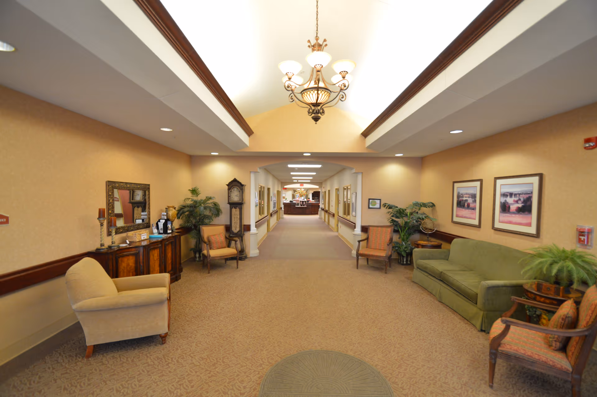 A well-lit hallway in a senior living facility with beige walls and carpet. The hallway features comfortable seating including armchairs and a green sofa, decorative plants, framed artwork on the walls, a wooden sideboard with a mirror, and a grandfather clock. A chandelier hangs from the ceiling, and the hallway leads to a reception or common area in the distance.
