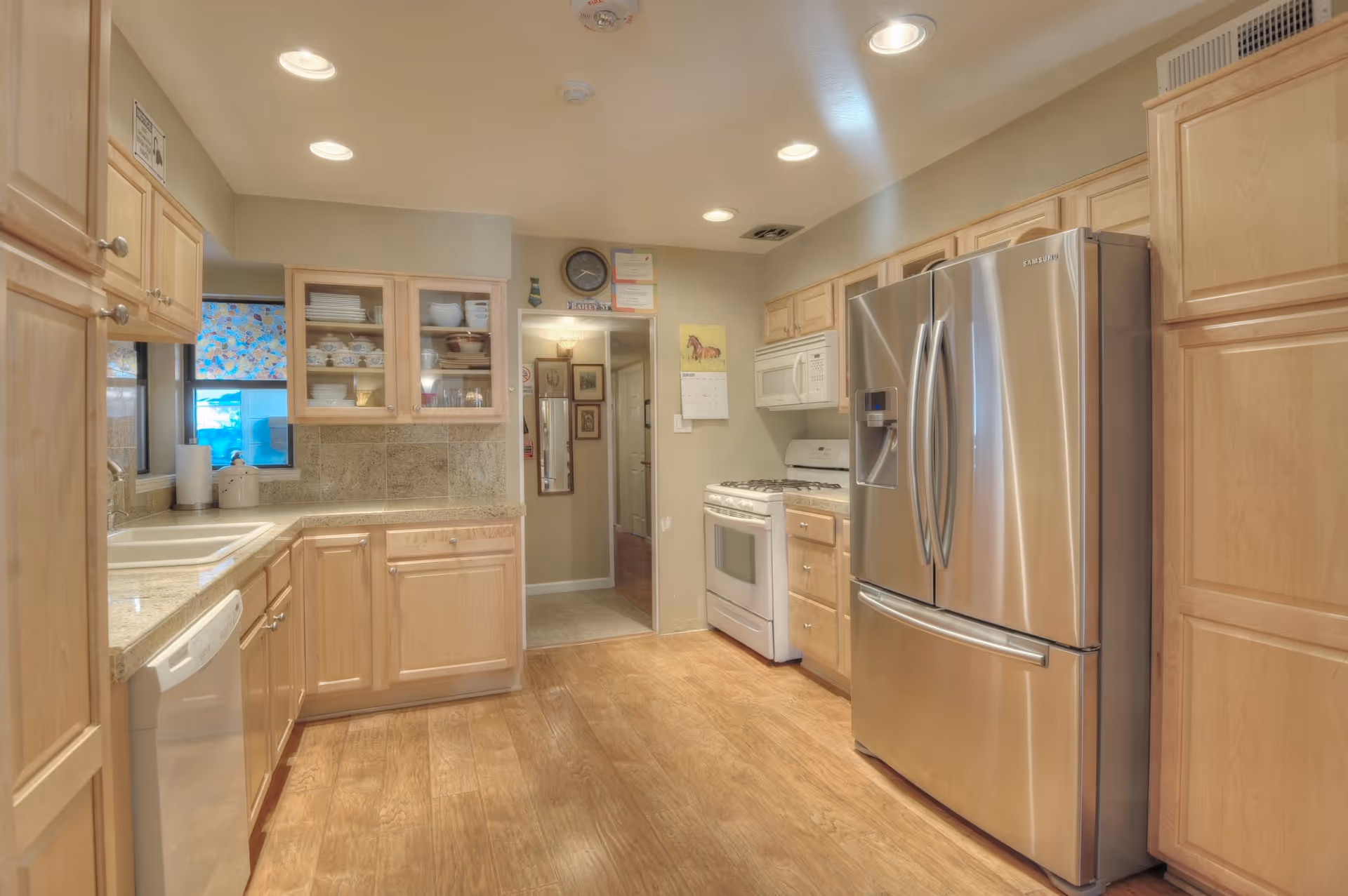 A bright kitchen with light wood cabinets, a stainless steel refrigerator, a white stove with a microwave above it, a dishwasher, and a sink under a window with a floral curtain. The floor is wooden and the ceiling has recessed lighting.