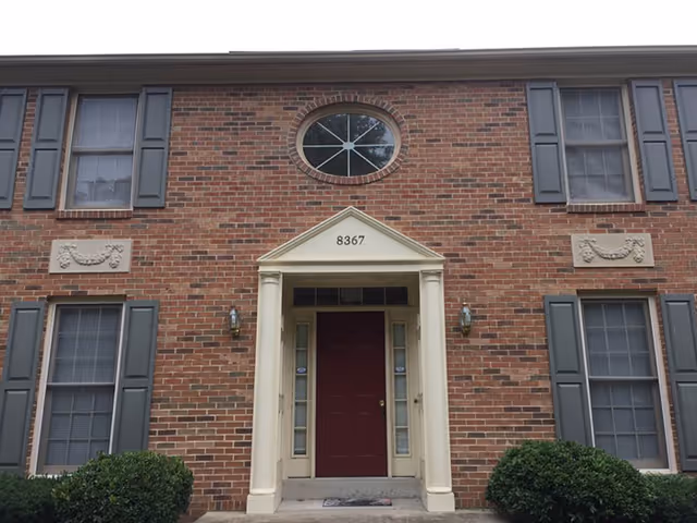 Front exterior view of a brick building with a maroon door centered under a small portico supported by two columns. The building has multiple windows with gray shutters and decorative stone accents. The address number 8367 is displayed above the door. Two bushes flank the entrance.