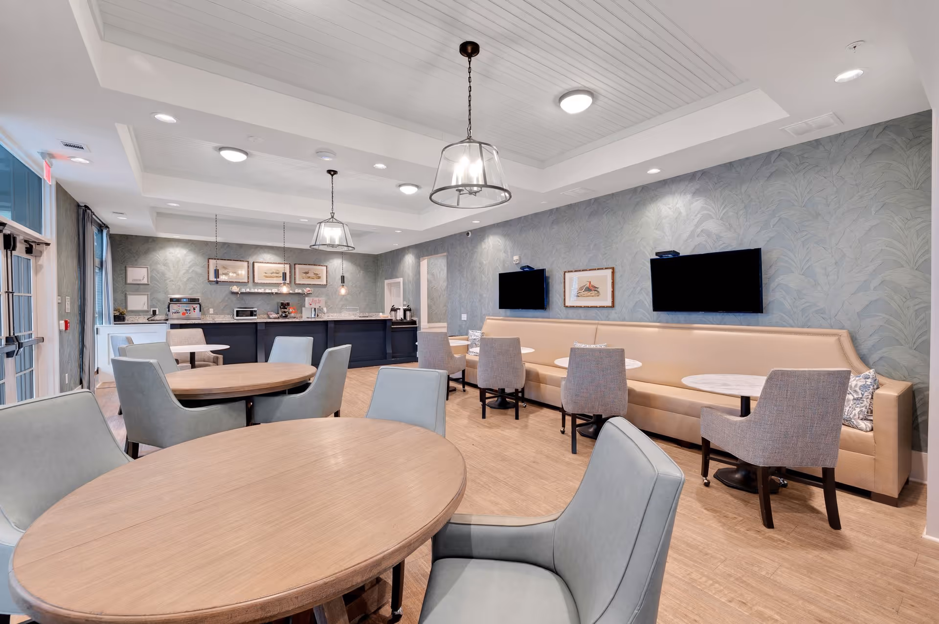 A bright and modern dining area with round wooden tables and cushioned chairs. The room features a long beige banquette along one wall with small round tables and additional chairs. Two flat-screen TVs and framed artwork are mounted on the wall above the banquette. Pendant lights hang from the white paneled ceiling, and a counter with coffee machines and other appliances is visible in the background. Large windows and a glass door allow natural light into the space.