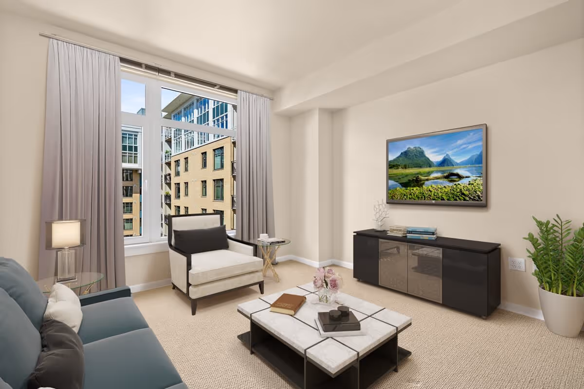 A bright living room with a large window showing an exterior view of adjacent buildings. The room features a blue sofa with pillows, a beige armchair with a black cushion, a glass side table with a lamp, a marble coffee table with books and a flower vase, a black TV stand with books and decorative items, a wall-mounted TV displaying a scenic landscape, and a potted plant in the corner.