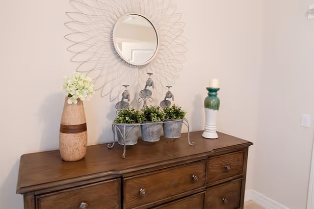 A wooden dresser with six drawers against a beige wall. On top of the dresser are decorative items including a tall vase with white flowers, a metal stand holding three small potted green plants, and a white and green candle holder with a white candle. Above the dresser is a round mirror with a decorative wire frame resembling a sunburst.
