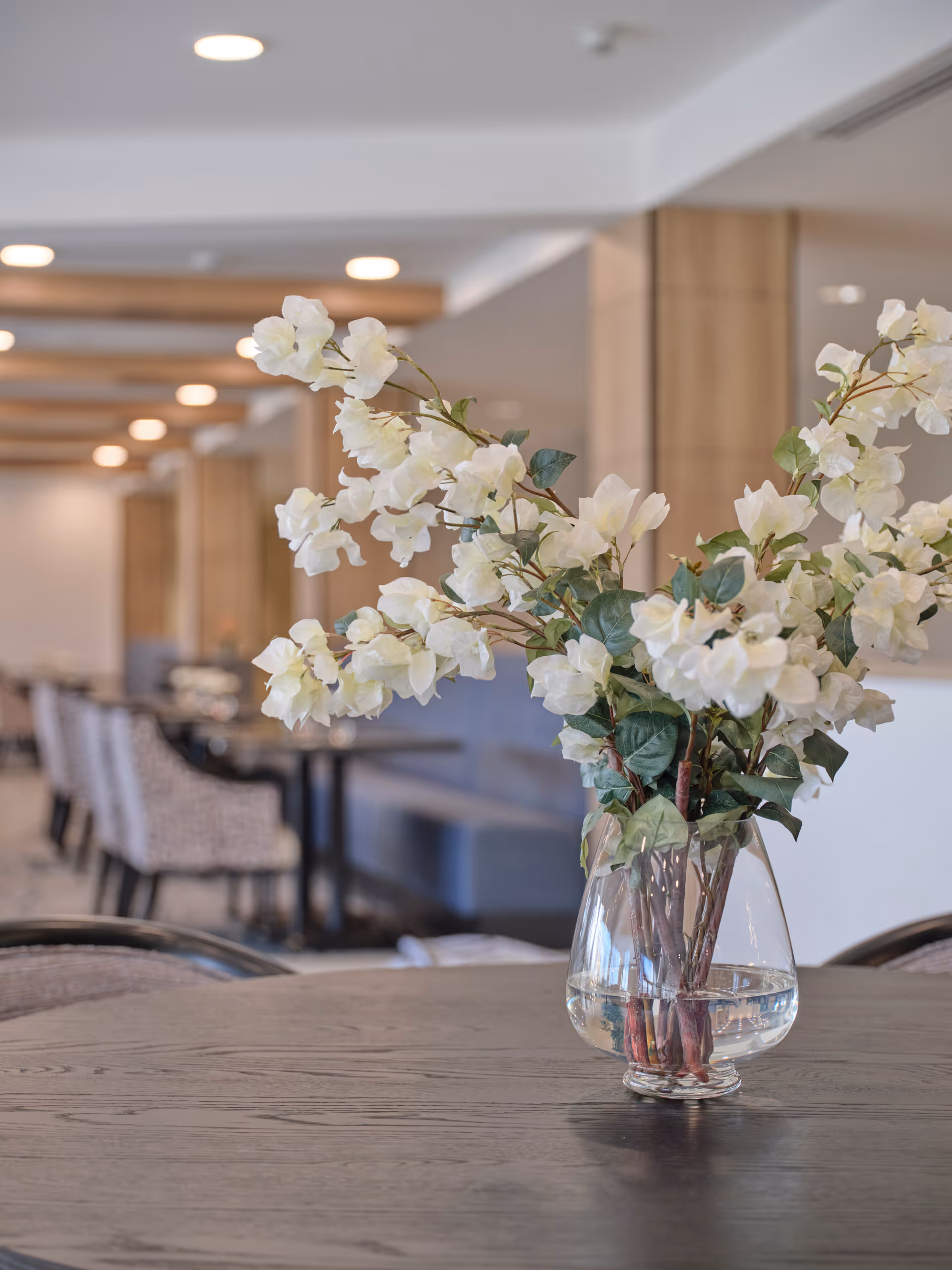 A clear glass vase with white flowers and green leaves sits on a dark wooden table in a well-lit dining area. In the background, there are upholstered chairs and tables arranged neatly, with wooden beams on the ceiling and soft lighting creating a warm atmosphere.