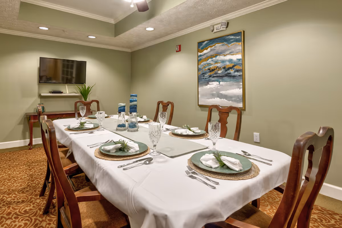 A dining room with a rectangular table set for six people. The table is covered with a white tablecloth and has green plates, folded white napkins, crystal glasses, and silverware. There are three blue decorative candles in the center. The room has light green walls, a patterned carpet, a wall-mounted TV, a small table with a plant, and a large abstract painting on the wall.