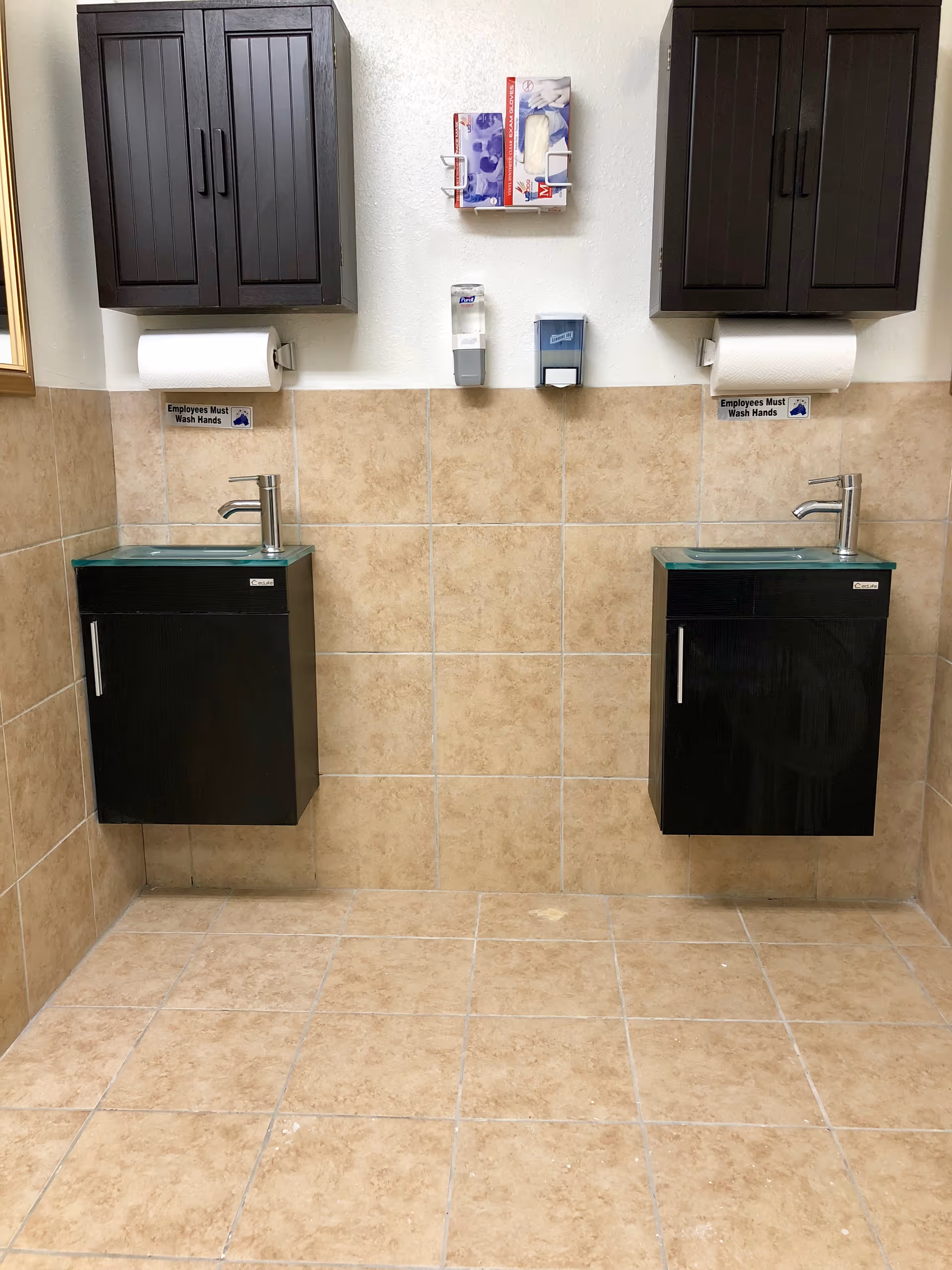 A clean restroom area with two black wall-mounted sinks with glass tops, each beneath a dark wooden cabinet. Above the sinks are paper towel holders, soap dispensers, and a wall-mounted box containing disposable gloves. The walls and floor are tiled in beige. Small signs above each sink read 'Employees Must Wash Hands.'