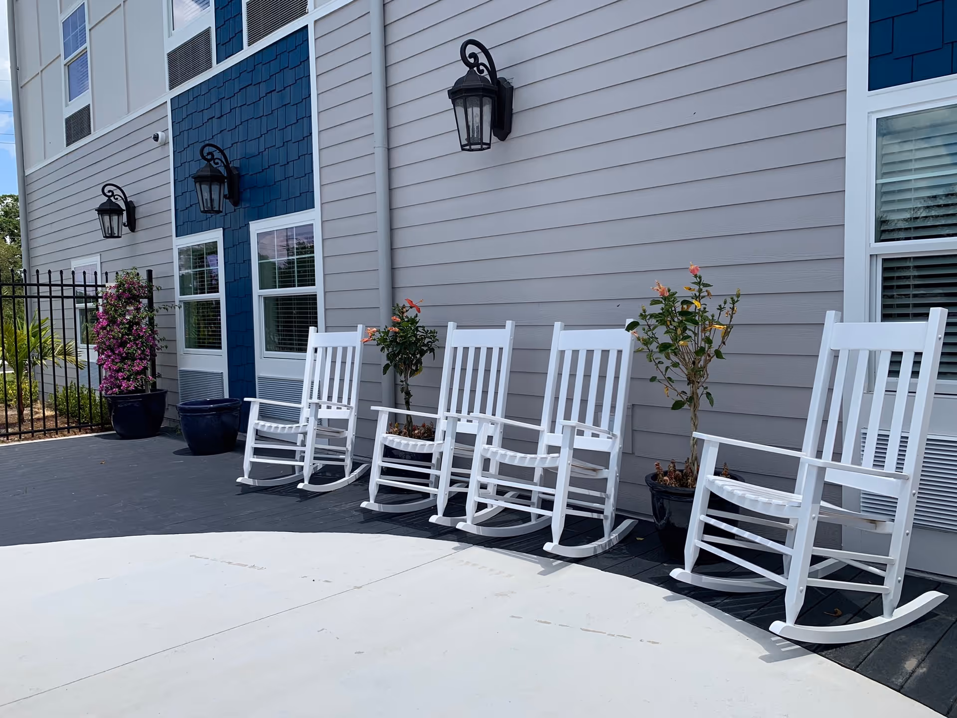 Outdoor patio area with five white rocking chairs lined up against the exterior wall of a building. The building has gray siding with a blue section and black wall-mounted lanterns. There are potted plants with flowers placed between the chairs and near a black metal fence.