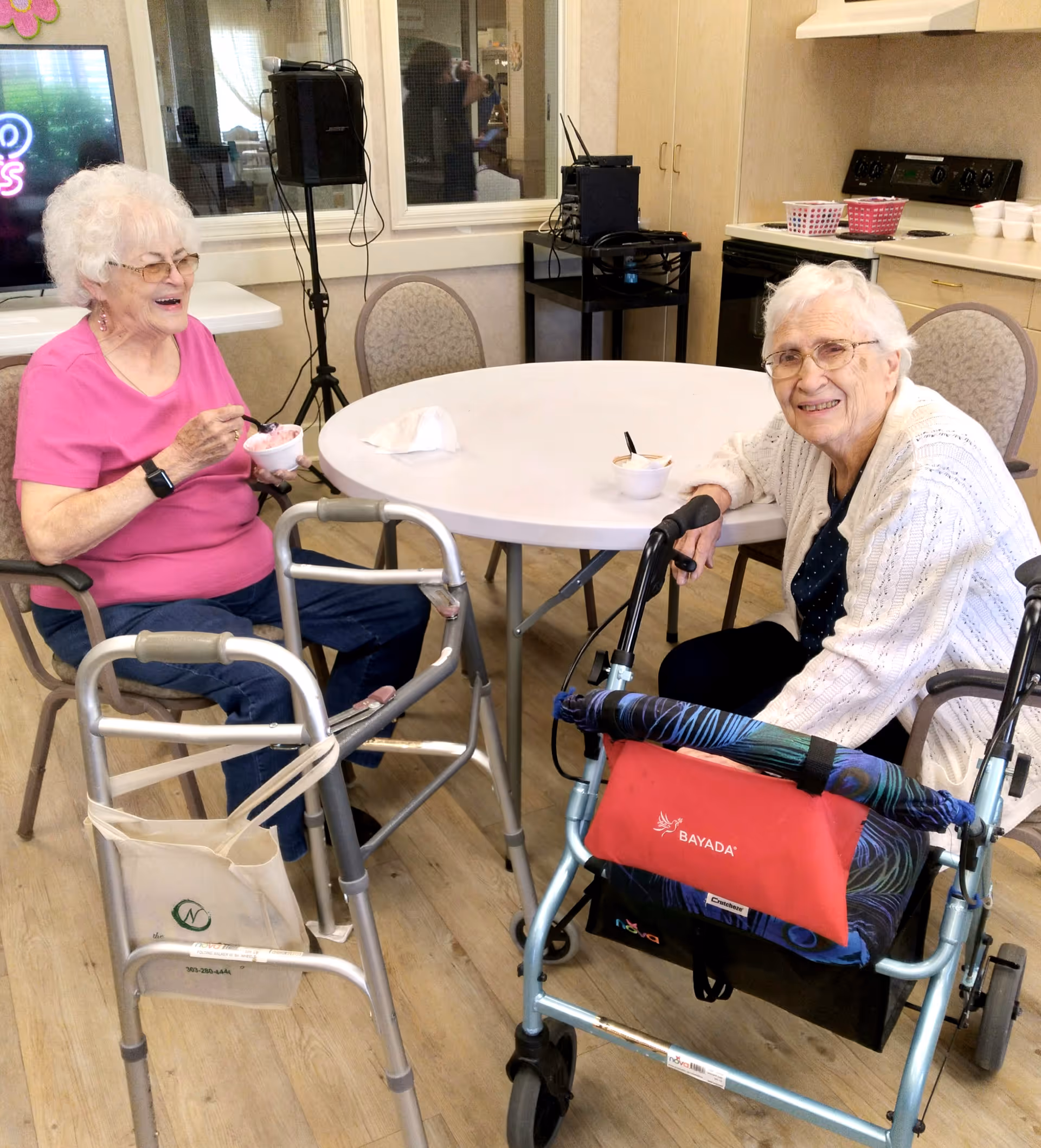 Two elderly women sitting at a round table in a senior living facility kitchen or dining area, enjoying bowls of food. Both women have walkers next to them. One woman is wearing a pink shirt and glasses, smiling while eating, and the other woman is wearing a white cardigan and glasses, smiling at the camera. The background shows kitchen cabinets, a stove, and some appliances.