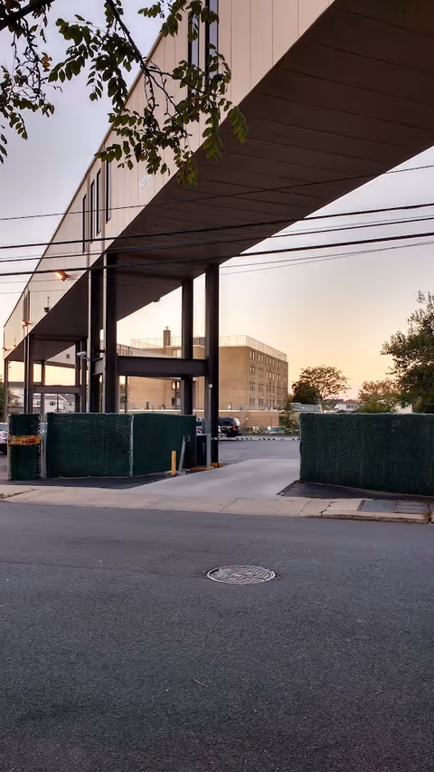 Entrance driveway beneath an elevated skybridge connecting two building sections, with parking, green temporary fencing, and a multi-story brick building in the background at sunset.