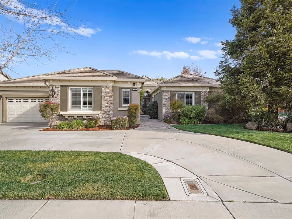 Front exterior view of a single-story residential building with stone and stucco facade, a two-car garage, a curved concrete driveway, well-maintained lawn, and landscaping including bushes and trees under a blue sky with scattered clouds.