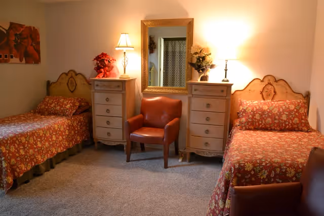 A cozy bedroom with two single beds, each covered with red floral bedspreads. Between the beds are two matching cream-colored dressers with lamps and flower arrangements on top. A brown leather armchair is positioned in the center beneath a large ornate mirror on the wall. The room has beige carpet and soft lighting.