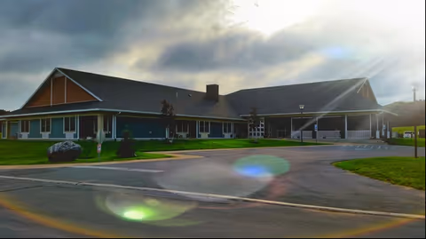 Single-story retirement community building with a covered entrance, driveway and lawn under a cloudy sky with sun flare.