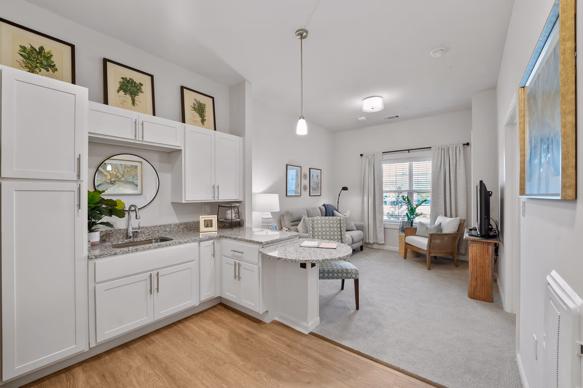 A bright and modern interior space featuring a small kitchen area with white cabinets, granite countertops, and a sink. Above the cabinets are three framed botanical prints. Adjacent to the kitchen is a cozy living room with a sofa, armchair, a window with curtains, a TV on a wooden stand, and framed artwork on the walls. The flooring transitions from wood in the kitchen to carpet in the living room.