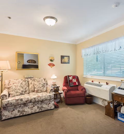 A cozy living room with a floral patterned sofa and a red armchair with a patchwork quilt draped over it. There is a small side table with a lamp between the sofa and armchair. A window with white blinds and a sheer valance lets in natural light. The walls are decorated with a mirror and small framed pictures. The floor is carpeted in beige.