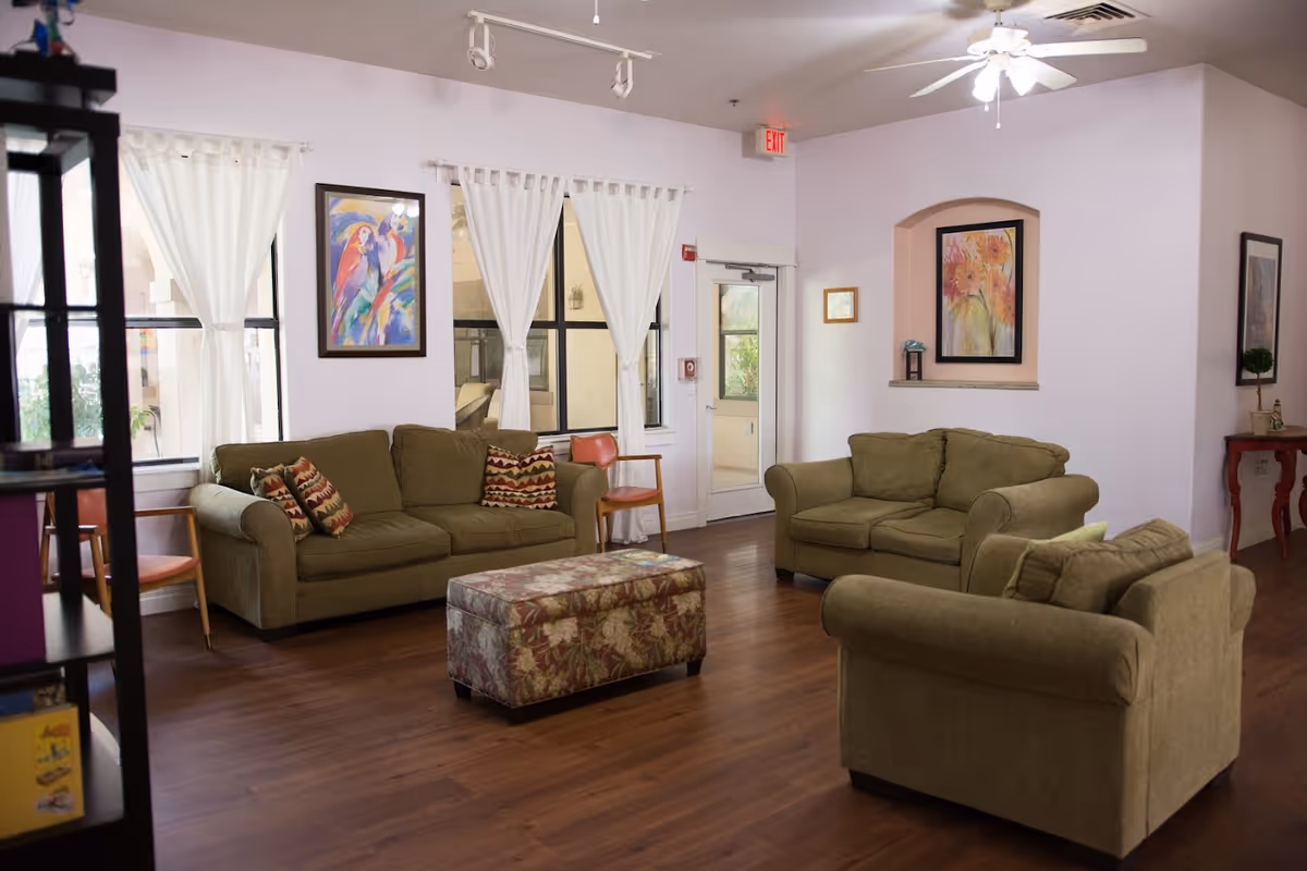 A cozy living room area with three olive green sofas arranged around a floral patterned ottoman. The room has wooden flooring, white walls, and three windows with white curtains. Colorful paintings hang on the walls, and there is a ceiling fan with lights. A door with a glass window leads outside, and there are a couple of wooden chairs and a small red table with decorative items.