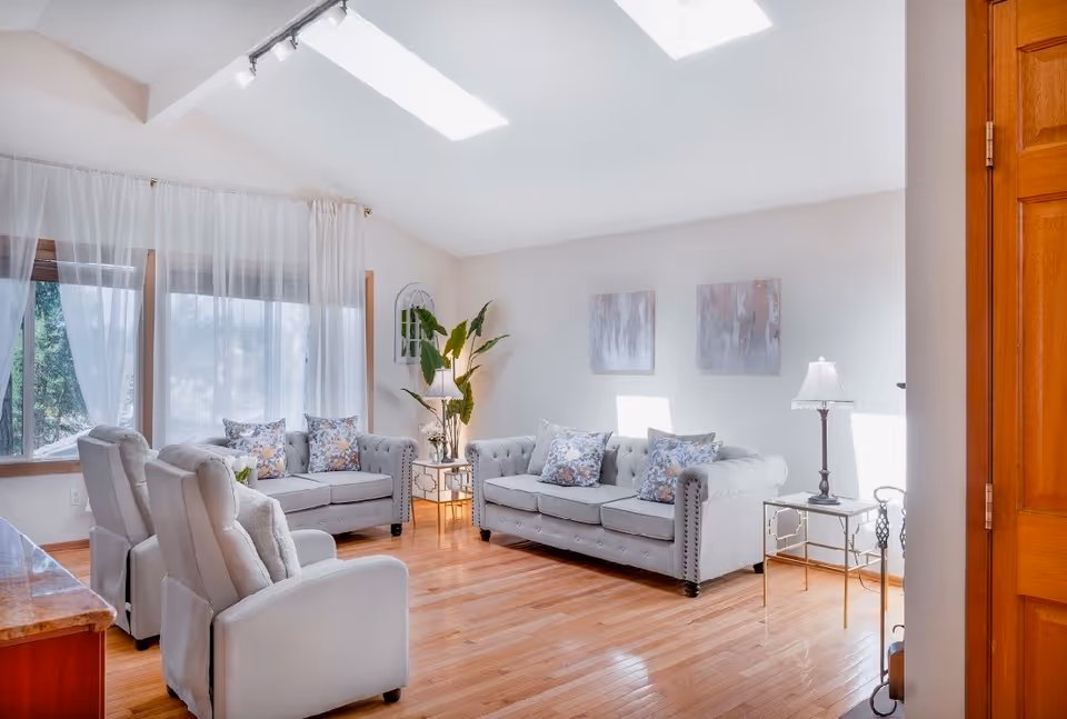Bright living room with gray sofas and armchairs, hardwood floors, large windows with sheer curtains, plants, and skylights.