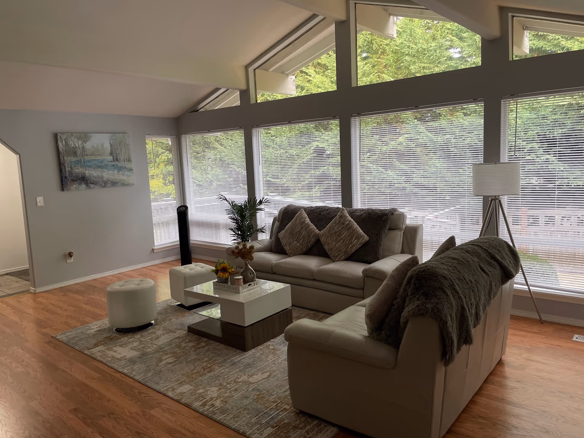 A cozy living room with large windows covered by blinds, letting in natural light and showing green trees outside. The room features two beige leather sofas with multiple cushions and a throw blanket, a modern coffee table with decorative items, a floor lamp with a tripod base, a small white ottoman, and a painting on the wall. The floor is wooden with a large area rug under the furniture.