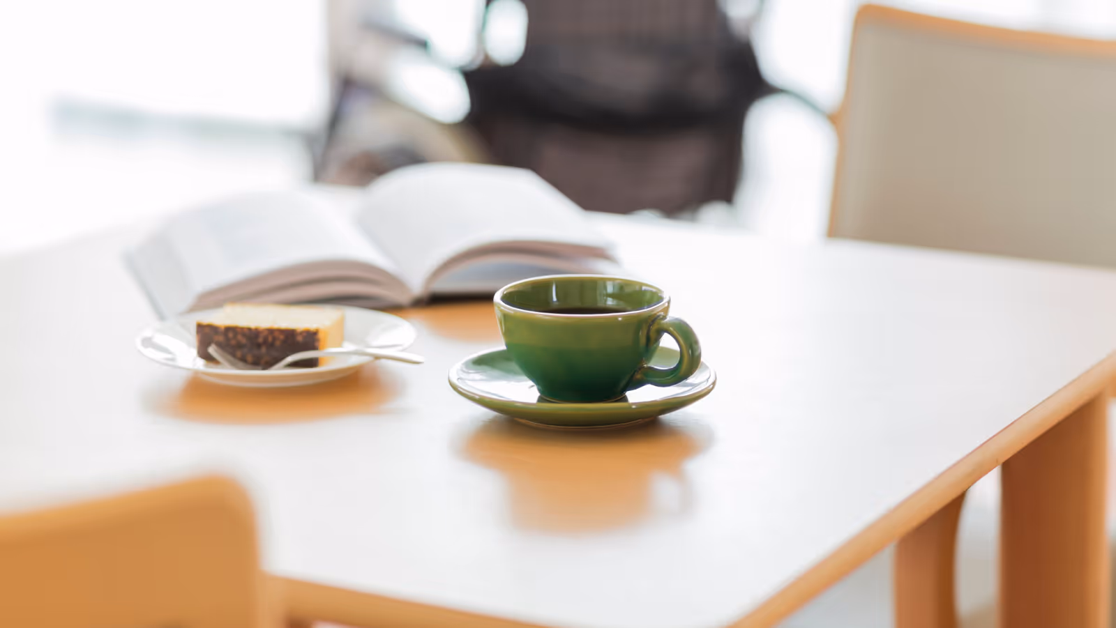 A green cup and saucer on a light wooden table with an open book and a plate holding a slice of cake in the background.
