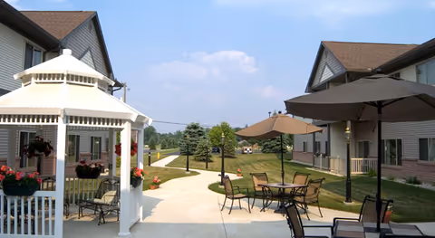 Outdoor patio area at Sweetwater Retirement Community featuring a white gazebo with hanging flower baskets, several tables with chairs and large umbrellas, surrounded by two-story residential buildings and a landscaped lawn with trees under a partly cloudy sky.