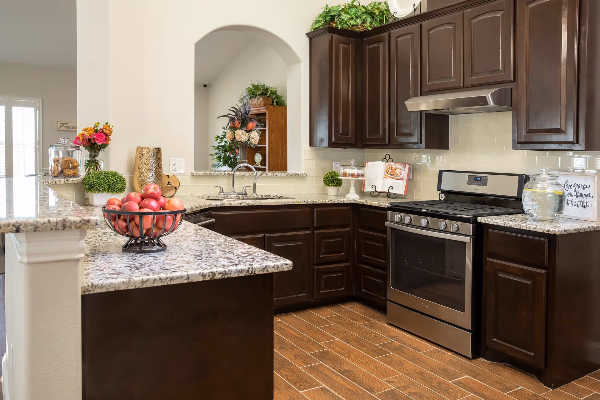 Modern kitchen with dark wooden cabinets, granite countertops, a stainless steel gas stove, and a double sink. The countertop has a bowl of red apples, a vase with flowers, a glass jar with cookies, and decorative plants. The floor is wooden with a tile pattern.