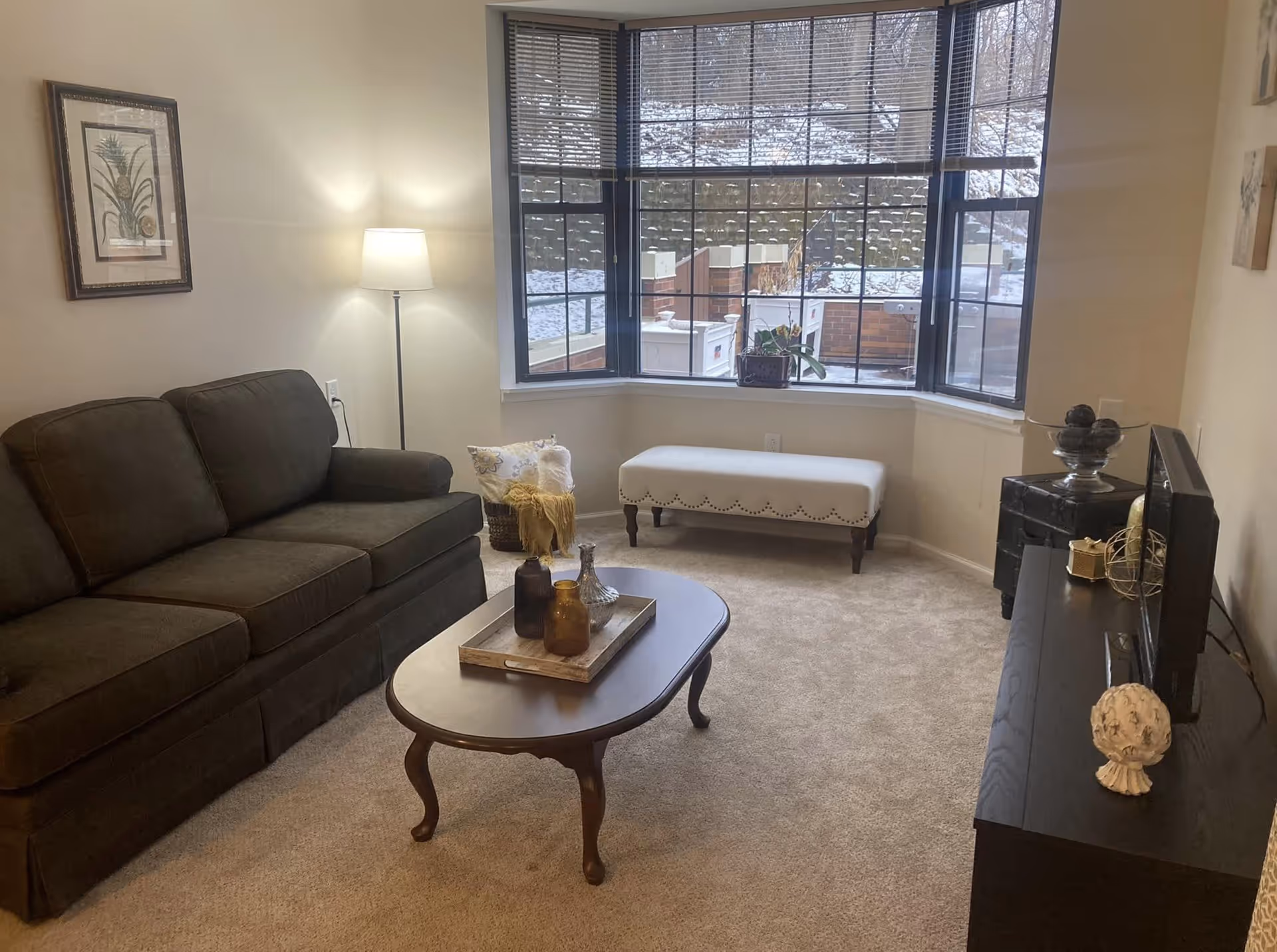 A cozy living room with a dark brown sofa on the left, a wooden coffee table with decorative bottles in the center, and a white cushioned bench under a large bay window with blinds. A floor lamp is lit next to the sofa, and a TV sits on a black stand on the right side of the room. The walls are light-colored, and there is a framed picture hanging above the sofa. Outside the window, snow is visible on the ground and brick structures.