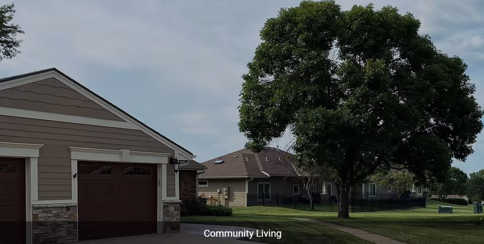 Exterior view of a residential community with a garage, a large tree, and multiple single-story houses surrounded by green lawns under a partly cloudy sky.