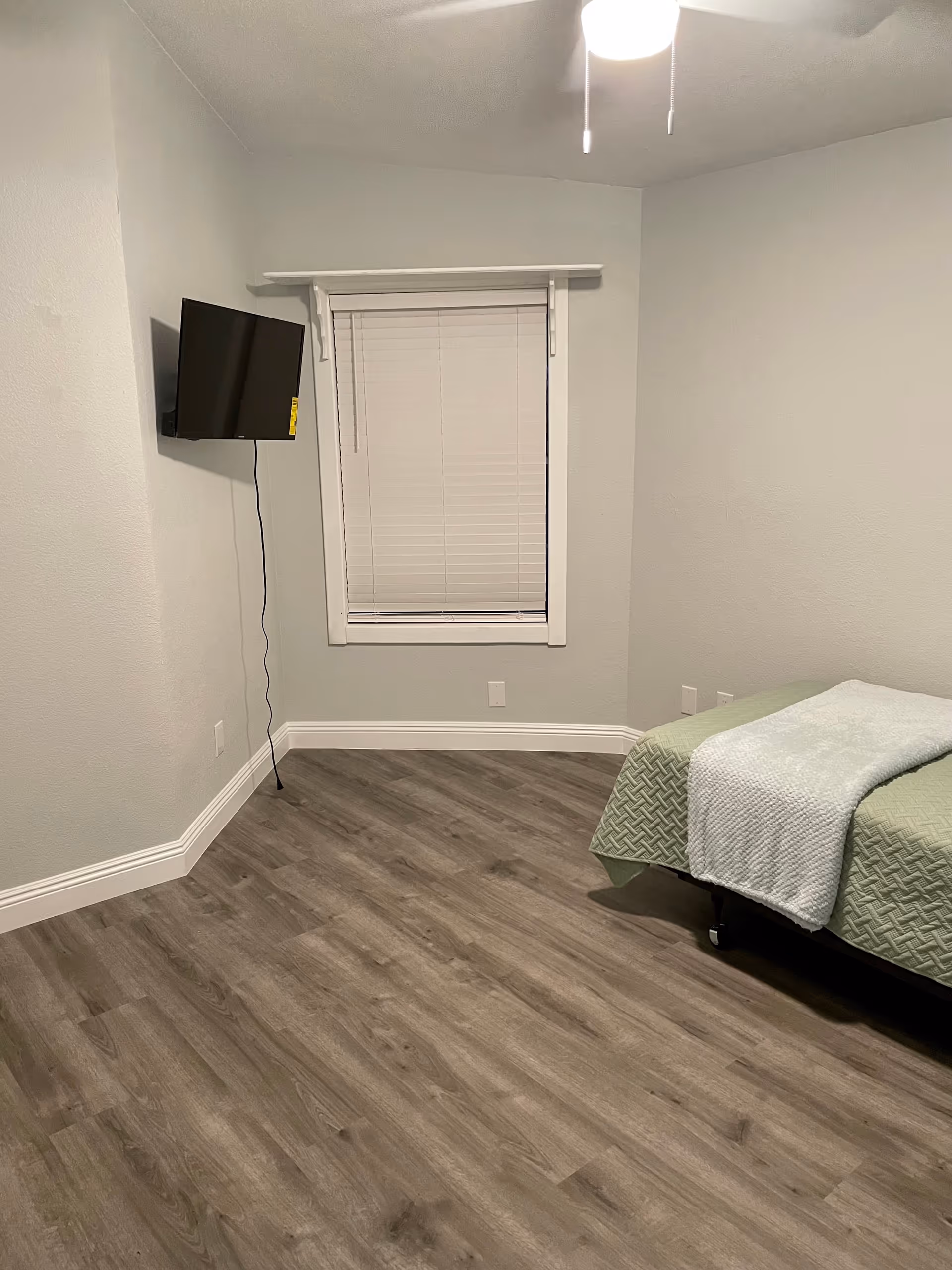 Small sparsely furnished bedroom with a wall-mounted TV, a window with blinds, wood-look flooring, and a bed with a green coverlet.