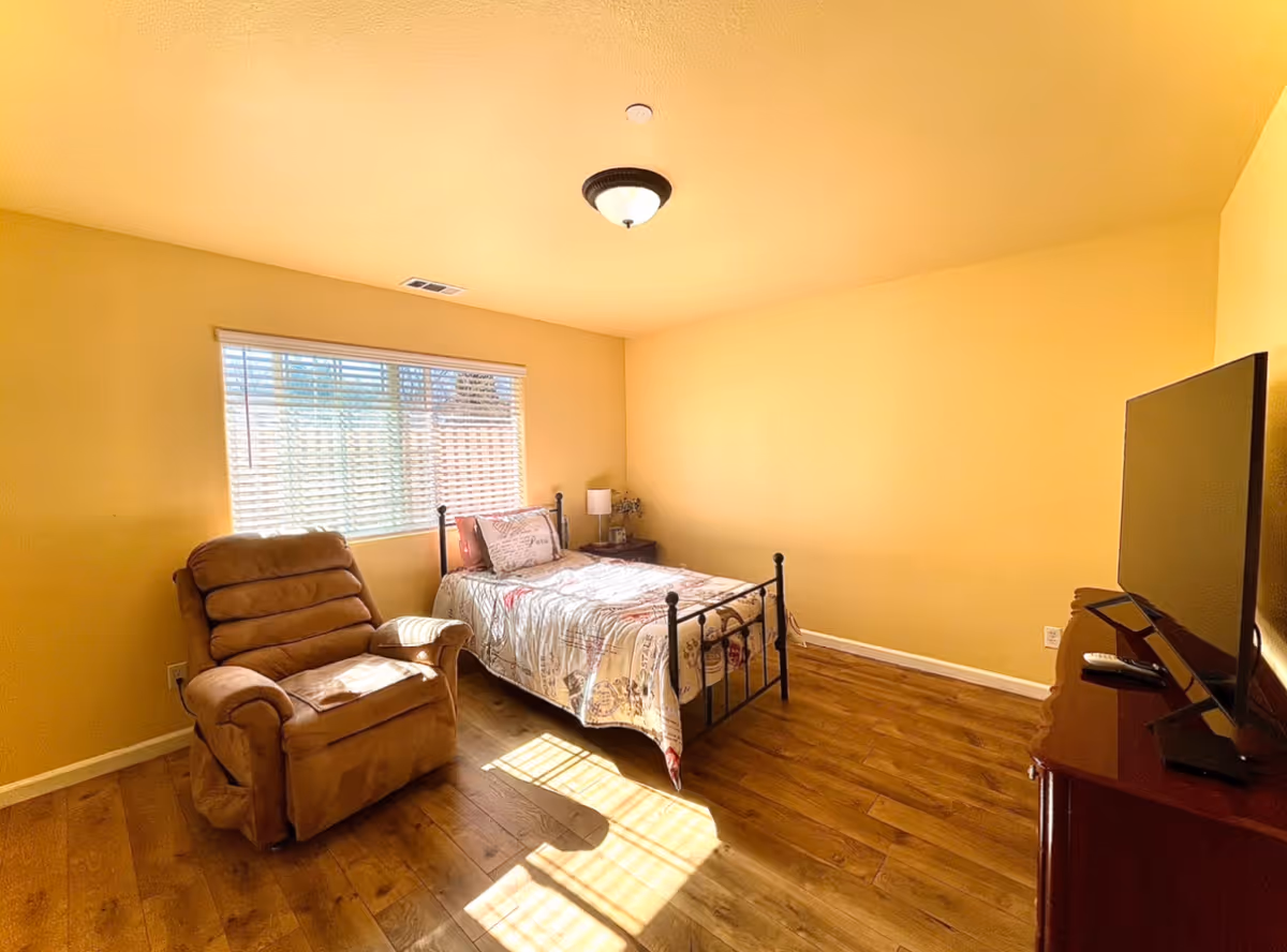 Sunlit bedroom with a single metal-framed bed, a tan recliner, a TV on a dresser, wood floors, and a window with blinds.