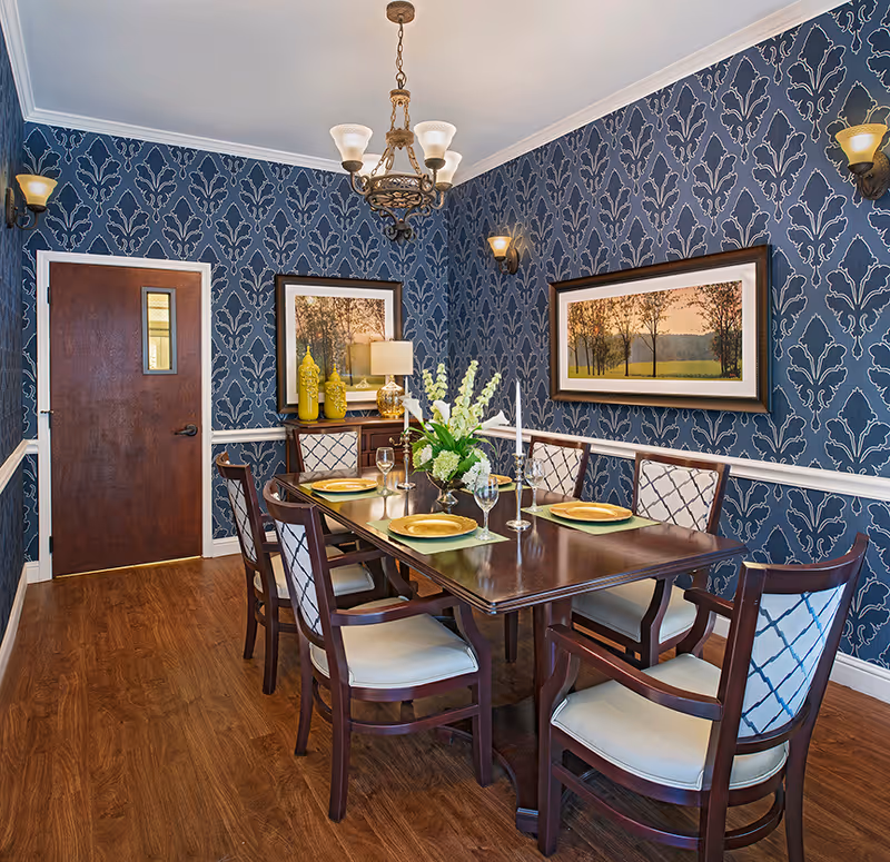 A formal dining room with a dark wooden table set for six people. The chairs have cushioned seats and backs with a patterned fabric. The walls are covered with blue wallpaper featuring a large floral pattern. Two framed landscape paintings hang on the walls. A chandelier and wall sconces provide lighting. A sideboard with decorative yellow vases and a lamp is against one wall. The floor is wood, and there is a wooden door with a small window.