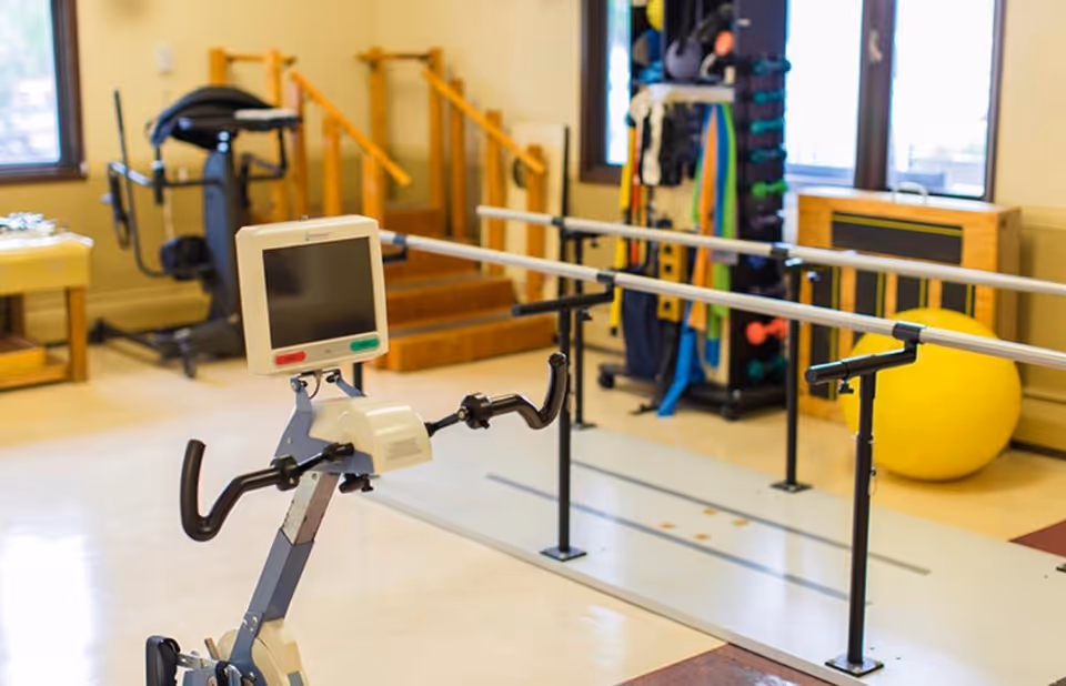 A rehabilitation room with exercise equipment including a stationary bike with a monitor, parallel bars for walking exercises, a yellow exercise ball, and various resistance bands and weights on a rack near windows.