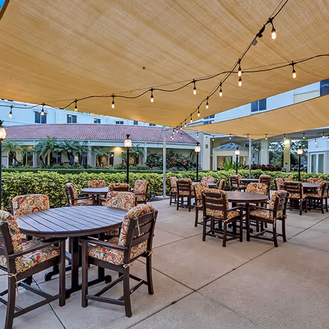 Outdoor patio area with multiple round tables and cushioned chairs under large beige shade sails. String lights are hanging above the seating area. In the background, there are green bushes, palm trees, and a building with a red-tiled roof and white walls.