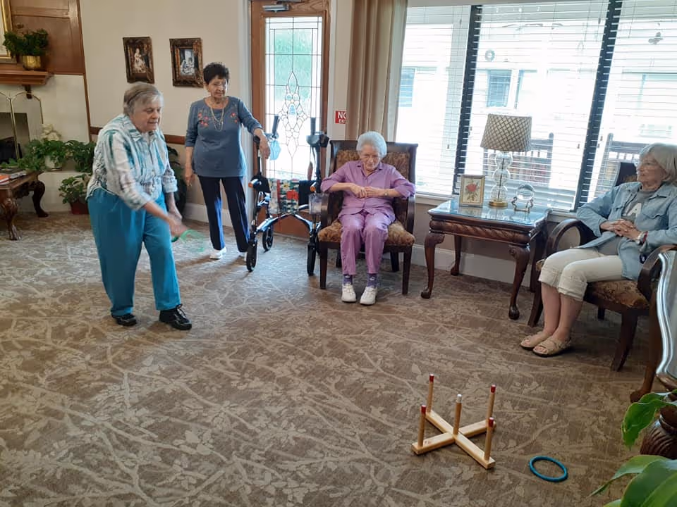 Four elderly women in a senior living facility common room playing a ring-toss game with chairs and a walker nearby.