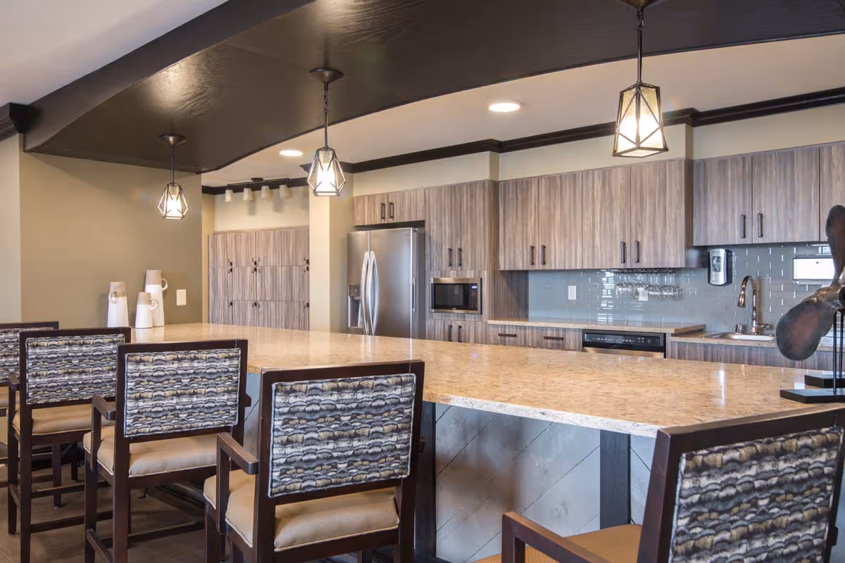 Modern kitchen area with a long marble countertop island surrounded by chairs with patterned backs. The kitchen features wood-finish cabinets, a stainless steel refrigerator, microwave, dishwasher, and a sink with a faucet. Pendant lights hang from the ceiling above the island.