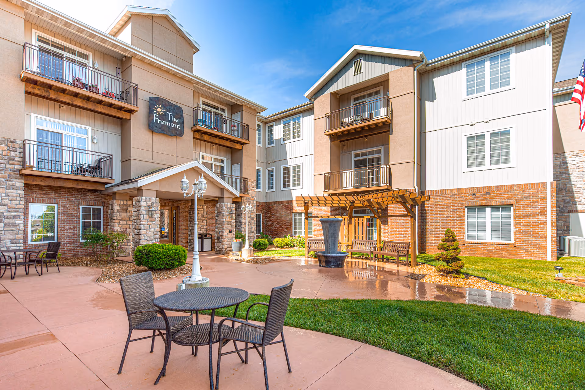 Exterior view of The Fremont Senior Living building showing a courtyard with patio tables, a fountain, balconies, and the main entrance.