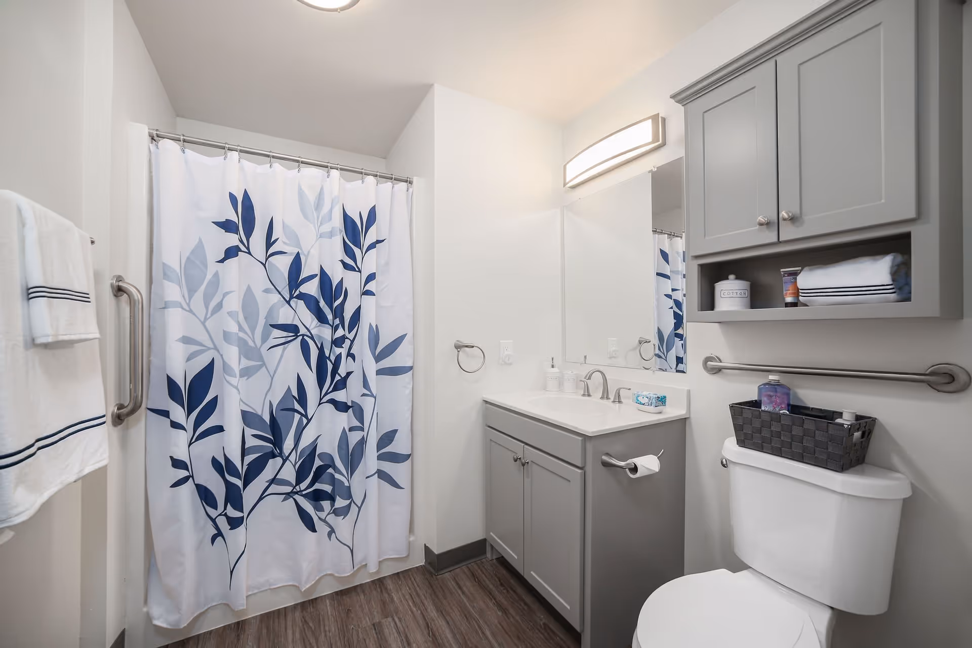 A clean and modern bathroom with a white toilet, a gray vanity with a white countertop and sink, a large mirror above the sink, and a wall-mounted cabinet with shelves holding towels and toiletries. There is a shower with a white curtain featuring a blue leaf pattern, a towel rack with a white towel, and a grab bar near the toilet.