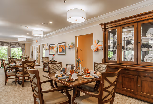 A well-lit dining room with wooden tables and chairs set for a meal, featuring plates of food, glasses of iced tea, and cups. The room has beige walls adorned with framed artwork and decorative wall hangings. A large wooden china cabinet displaying plates and glassware is visible on the right side. Large windows with curtains allow natural light to enter the space.